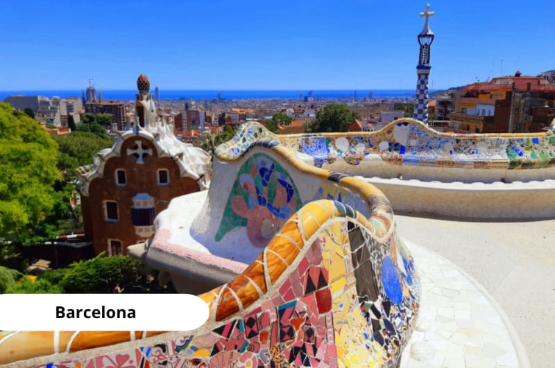 Vista panorámica del Parque Güell en Barcelona, con sus coloridos mosaicos, formas onduladas y arquitectura modernista diseñada por Gaudí, rodeado de vegetación y con la ciudad extendiéndose al fondo.