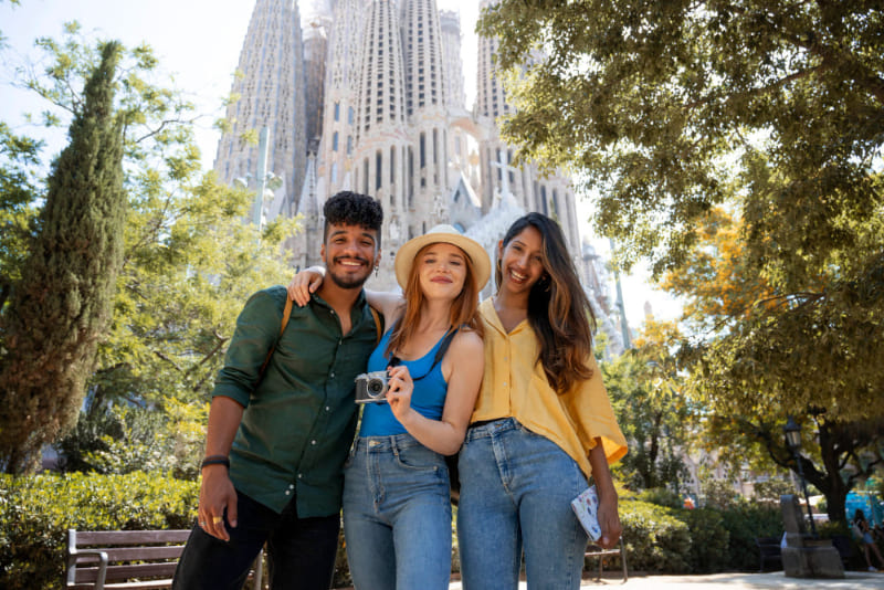 Tres turistas posan sonrientes frente a la Sagrada Familia en Barcelona, disfrutando de una visita turística en un día soleado.