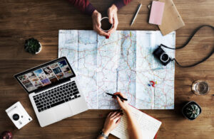 Manos planificando un viaje con un mapa, una cámara fotográfica y un portátil sobre una mesa.