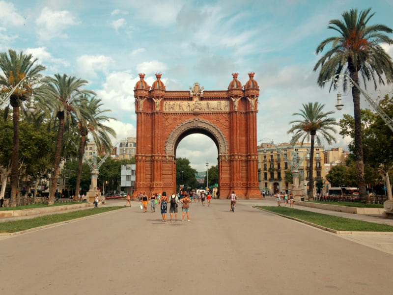 Vista del Arco del Triunfo de Barcelona enmarcado por una amplia avenida peatonal, con palmeras a los lados y personas paseando.