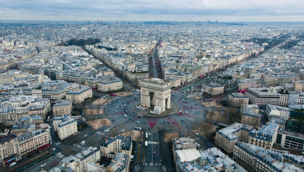Vista aérea del Arco del Triunfo en París, rodeado por la plaza Charles de Gaulle y las avenidas que convergen en el monumento, con la ciudad extendiéndose hasta el horizonte bajo un cielo nublado.