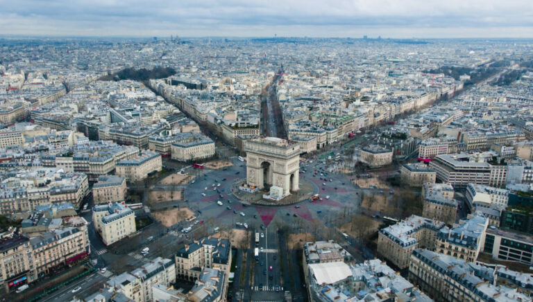 Vista aérea del Arco del Triunfo en París, rodeado por la plaza Charles de Gaulle y las avenidas que convergen en el monumento, con la ciudad extendiéndose hasta el horizonte bajo un cielo nublado.