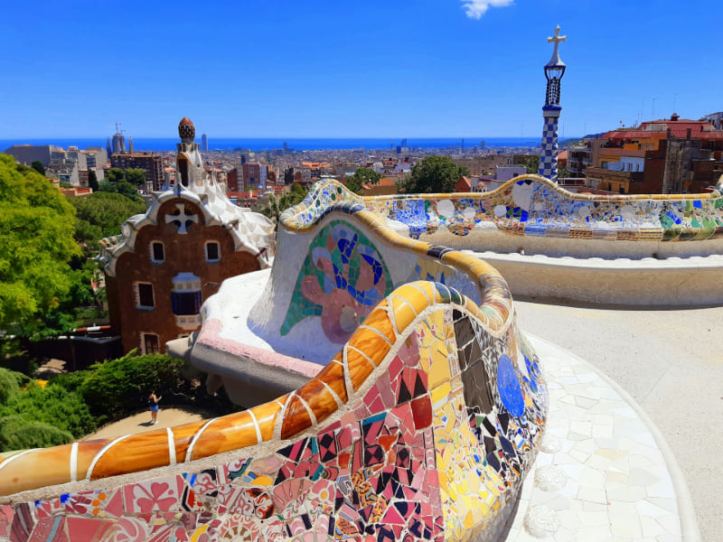 Vista panorámica del Parque Güell en Barcelona, con sus coloridos mosaicos, formas onduladas y arquitectura modernista diseñada por Gaudí, rodeado de vegetación y con la ciudad extendiéndose al fondo.