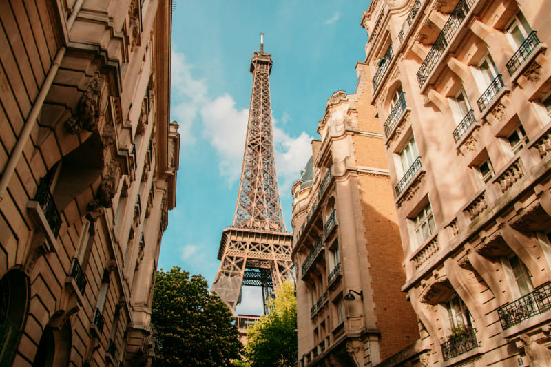 Vista de la Torre Eiffel desde una calle parisina, rodeada de edificios históricos y bajo un cielo azul parcialmente despejado.